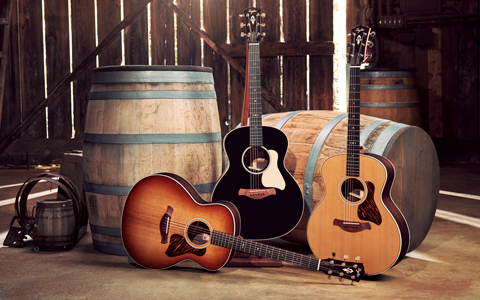 Selection de differentes guitares acoustiques de la Taylor Gold Label Collection posées sur le sol d une cabane en bois avec de vieux tonneaux de vin en arriere-plan